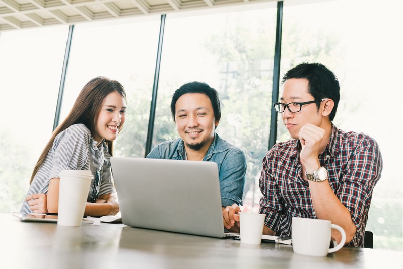 Group of young Asian business colleagues or college students using laptop in team casual discussion, startup project meeting or happy teamwork brainstorm concept, at coffee shop or modern office